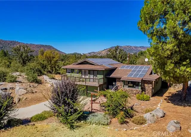 Two-story Ahwahnee home with a stone-accented facade, rooftop solar panels and a wraparound balcony, set among granite boulders and pine trees with forested hills under a clear blue sky