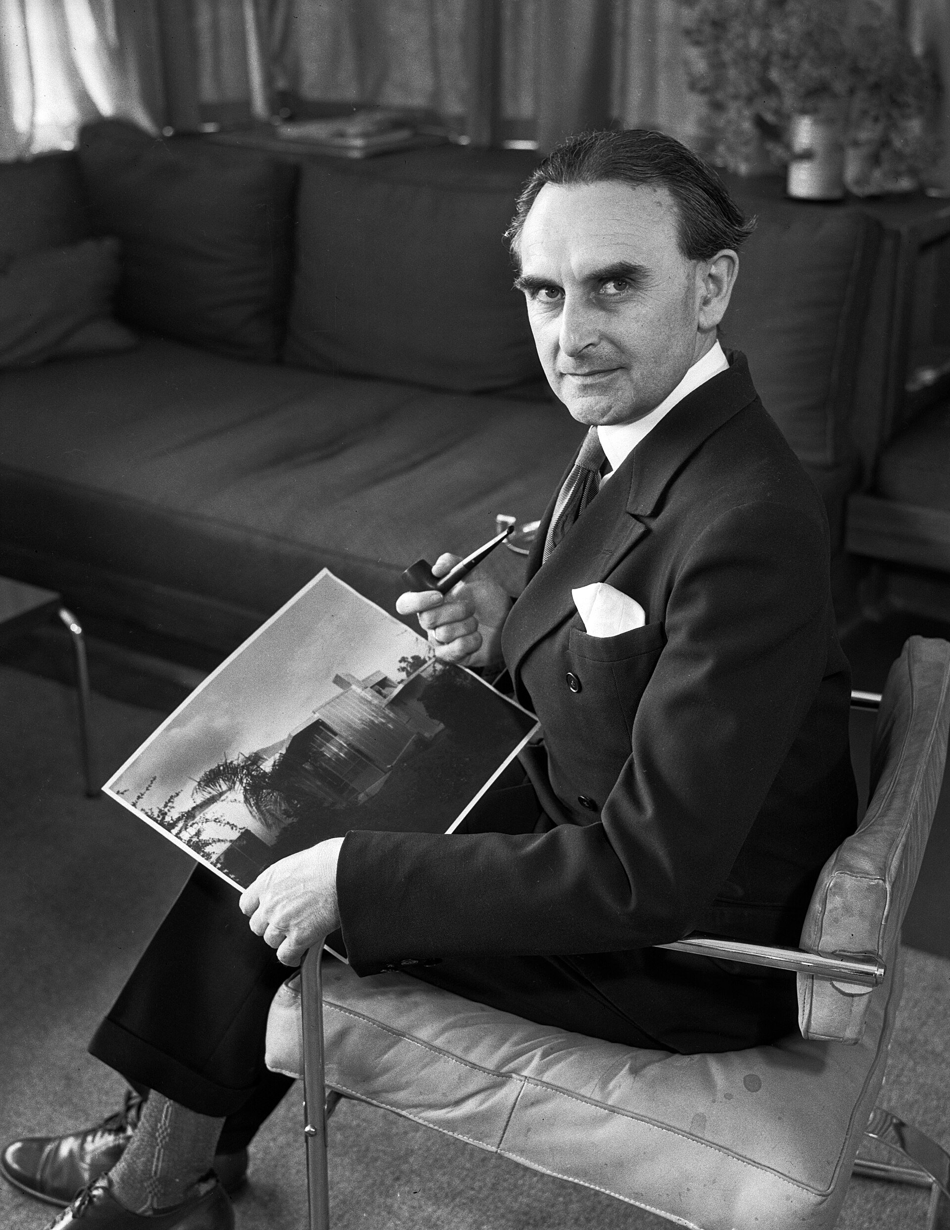 Richard Neutra seated on a cantilever chair of his own design, holding a pipe and a photograph of the Beard House.