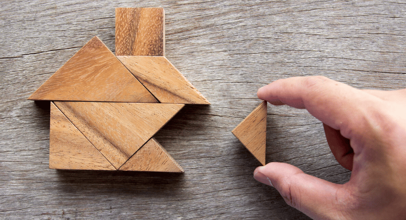 A hand places a wooden puzzle piece beside a house-shaped tangram on a wood surface.
