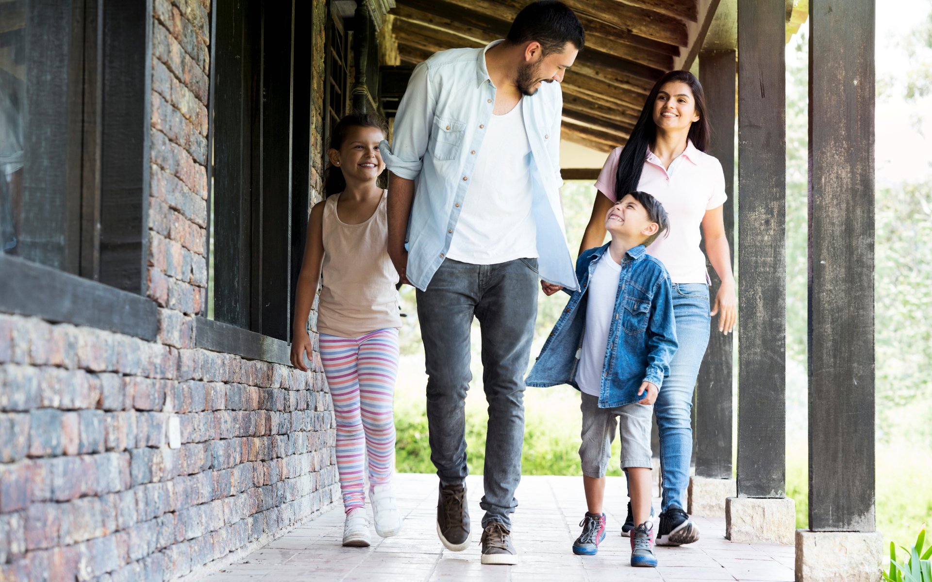 A family of four walks together along a covered stone-and-wood porch.