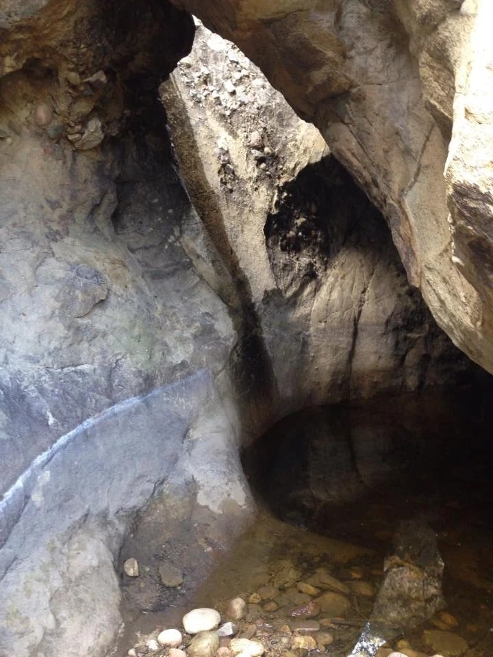 Rocky cave interior with a dark recess and shallow water over scattered stones.