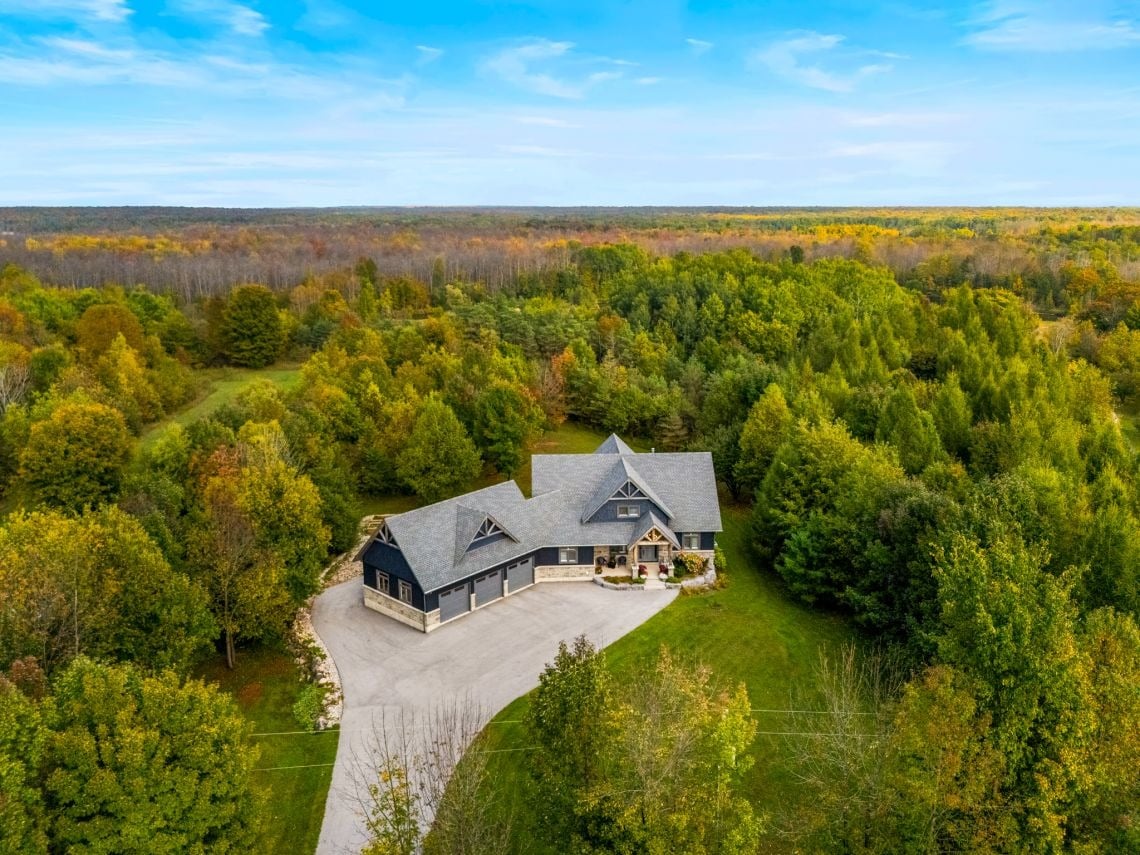 Aerial view of a large home amid autumn woodland, with a curved driveway and broad lawn.