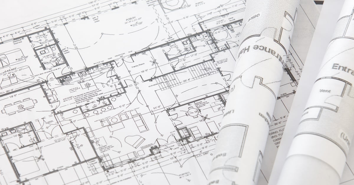 A close-up of architectural plans on for a large house on a table with two other, rolled up, plans lay on top of it.