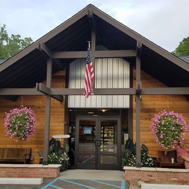 Wood-sided storefront with a timber gabled entrance, American flag, and hanging baskets of pink flowers.