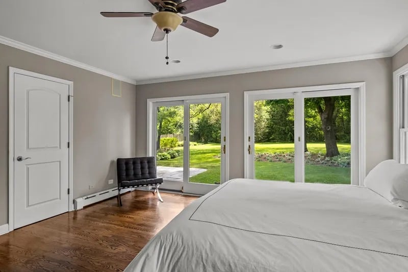 Bedroom with hardwood floors, a ceiling fan, and sliding glass doors overlooking a leafy lawn.