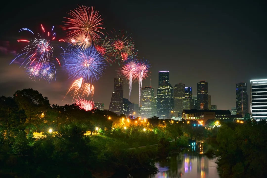 Houston skyline beneath colorful fireworks, reflected in water beside a tree-lined park.