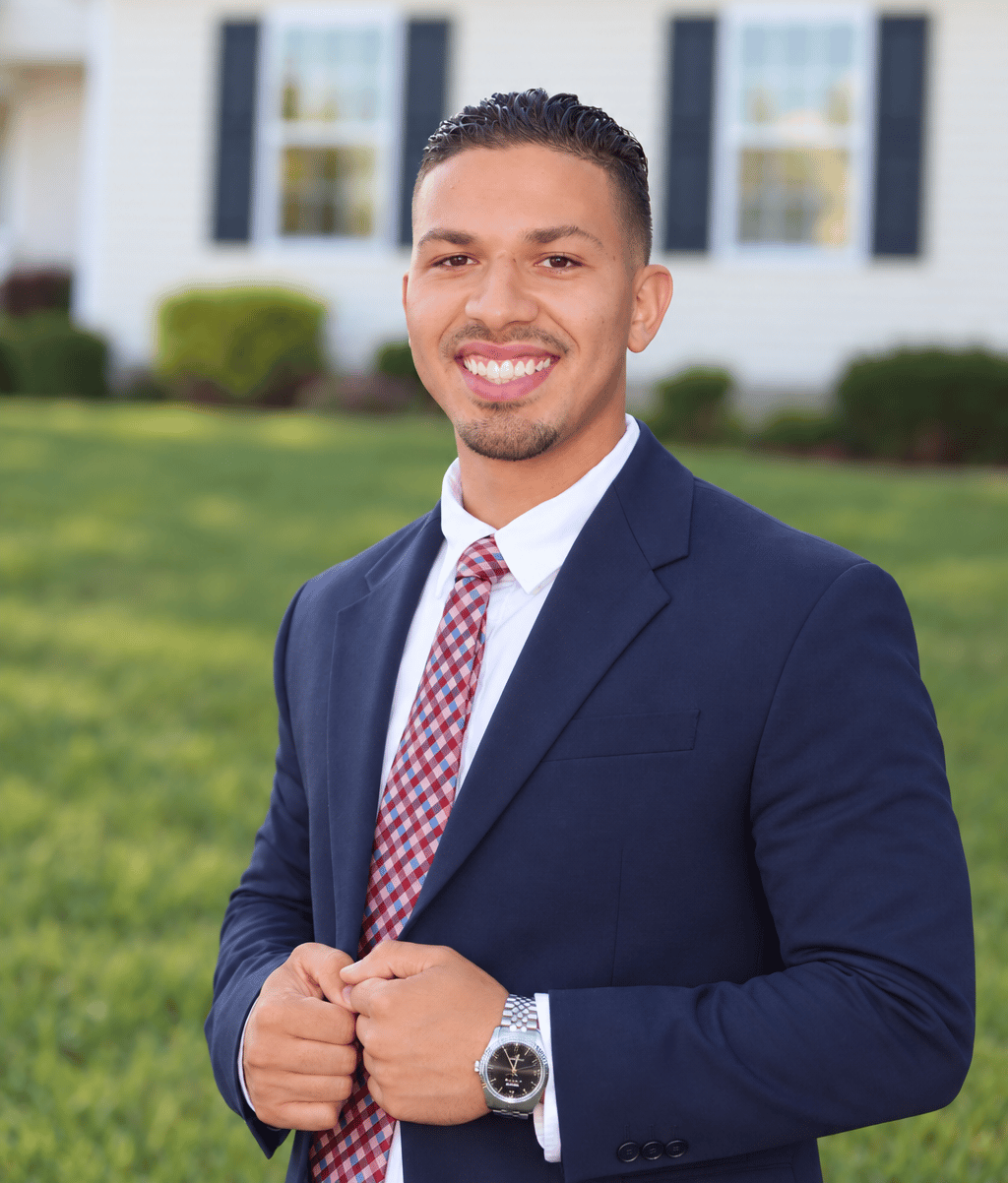 Anthony Bisono in a navy suit and patterned tie, standing in front of a house.