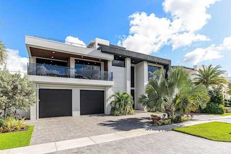 Modern two-story home with a glass balcony, dark garage doors, palms, and a paved driveway.