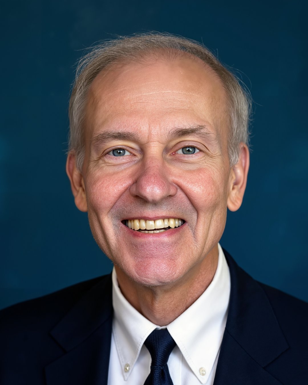 Bill Keim smiling in a dark suit, white shirt, and tie against a blue background.