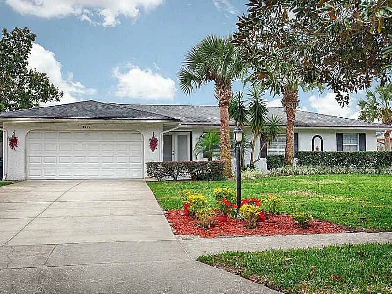 Single-story white house with an attached two-car garage, tropical palms, and a flower bed by the driveway.