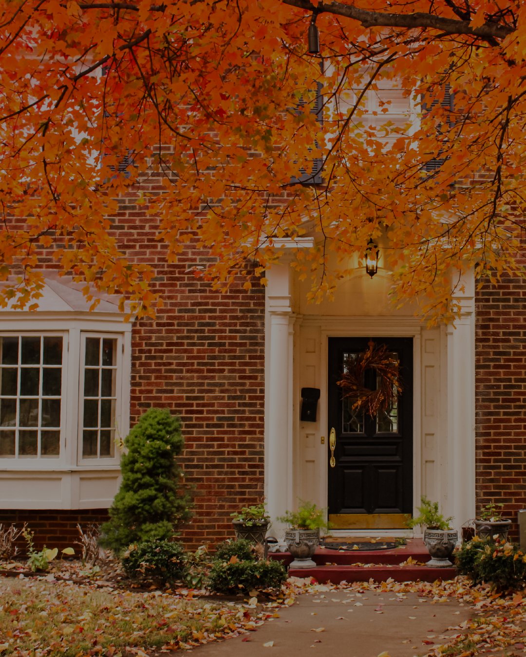 Brick home entrance with a white portico and black wreath-adorned door beneath orange autumn leaves.