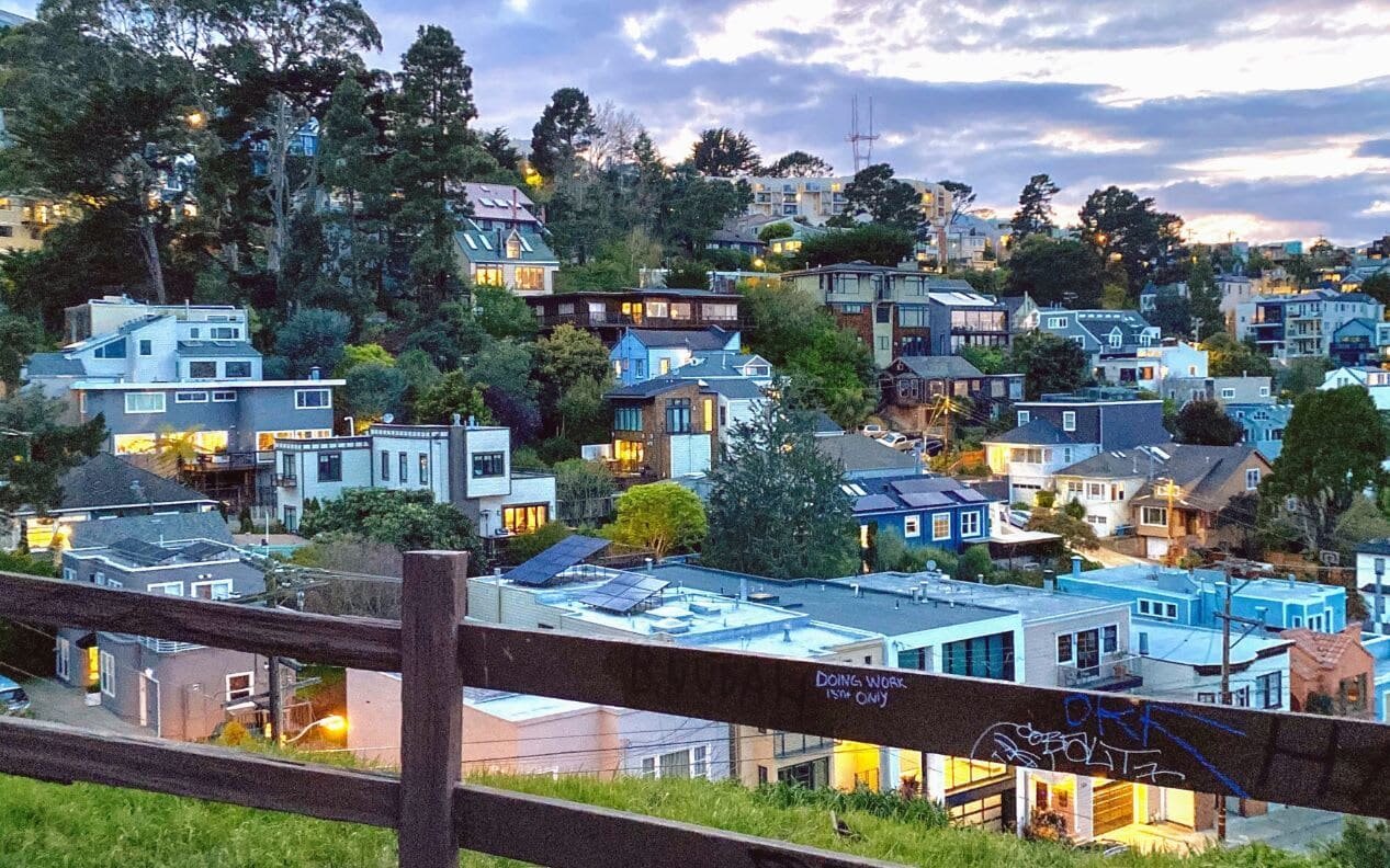 Noe Valley neighborhood in San Francisco, featuring a street lined with colorful Victorian and Edwardian homes, tree-lined sidewalks, and a backdrop of rolling hills under a clear blue sky.