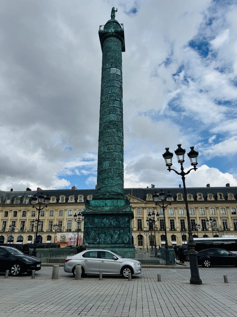 The Vendôme Column rises above parked cars and ornate Parisian buildings under a cloudy sky.