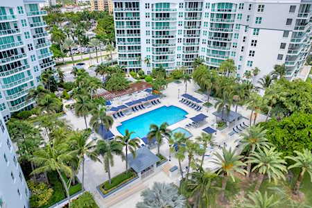 Aerial view of white condominium buildings surrounding a pool and palm-lined walkways.