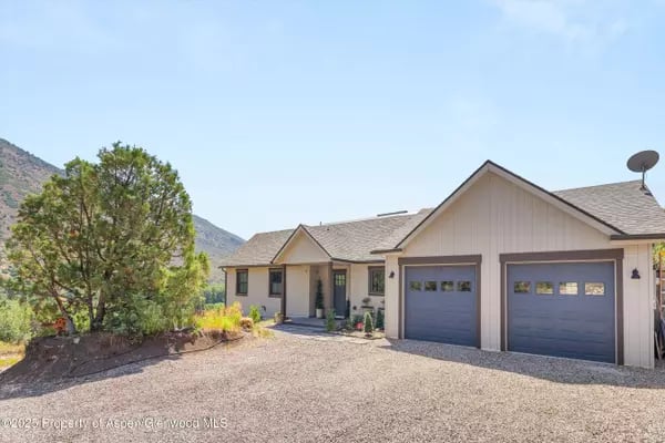House with two blue-gray garage doors, a gravel driveway, and mountain slopes in the background.