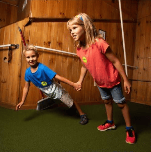 Two children lean together on a sloping floor inside a wooden room.