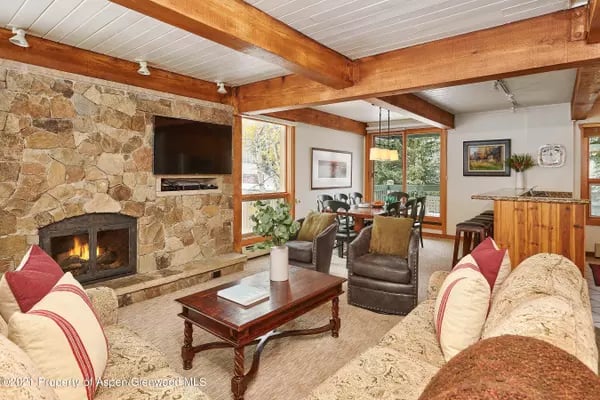 Living room with a stone fireplace, exposed timber beams, and dining area by tall windows.