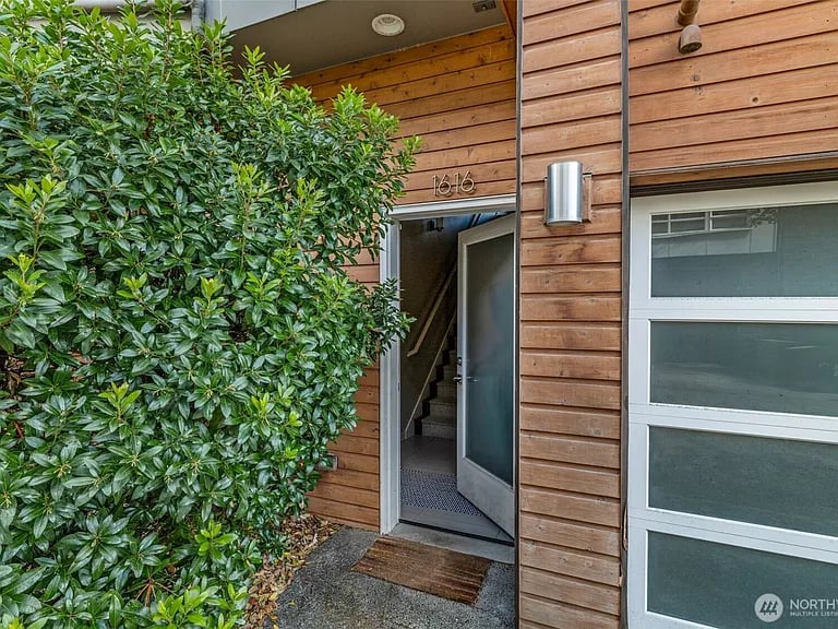 Wood-sided property entrance with an open door, number 1616, and a large shrub.
