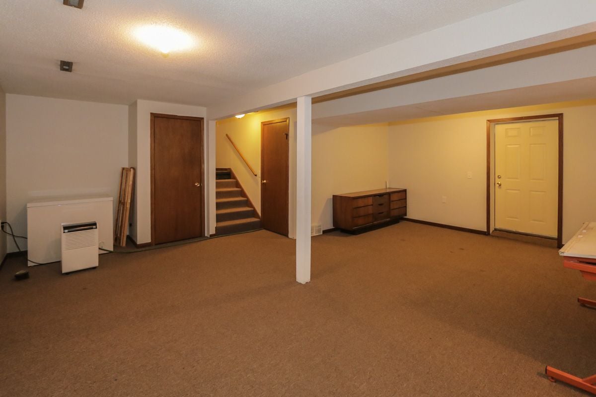 Basement room with brown carpet, a central support post, stairs, and a wooden dresser.