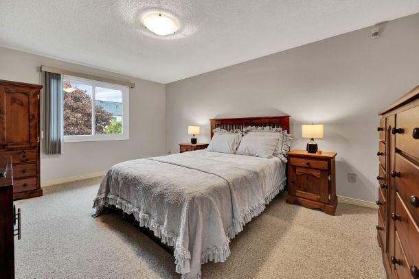 Bedroom with a gray-dressed bed, wooden furniture, beige carpet, and a window overlooking trees.