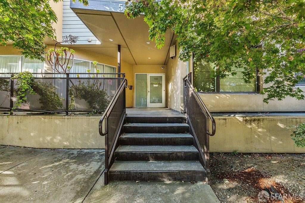 Residential building entrance with concrete steps, black railings, and a glass door beneath leafy trees.