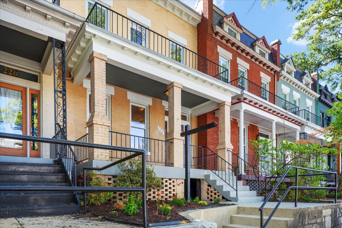 Pale yellow brick rowhouse with a covered porch, black railings, front steps, and a small garden.