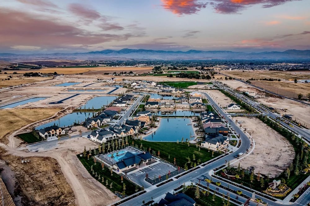 Aerial view of homes around a central pond, with construction areas and mountains in the distance.