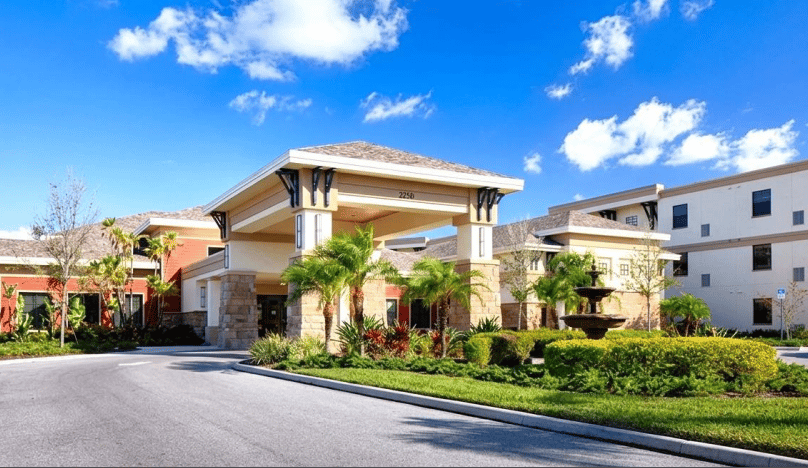 Senior living community entrance with a covered drive, palm trees, and a landscaped fountain.