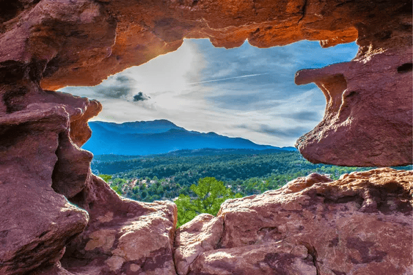 Forested foothills and distant mountains seen through a red sandstone rock opening.