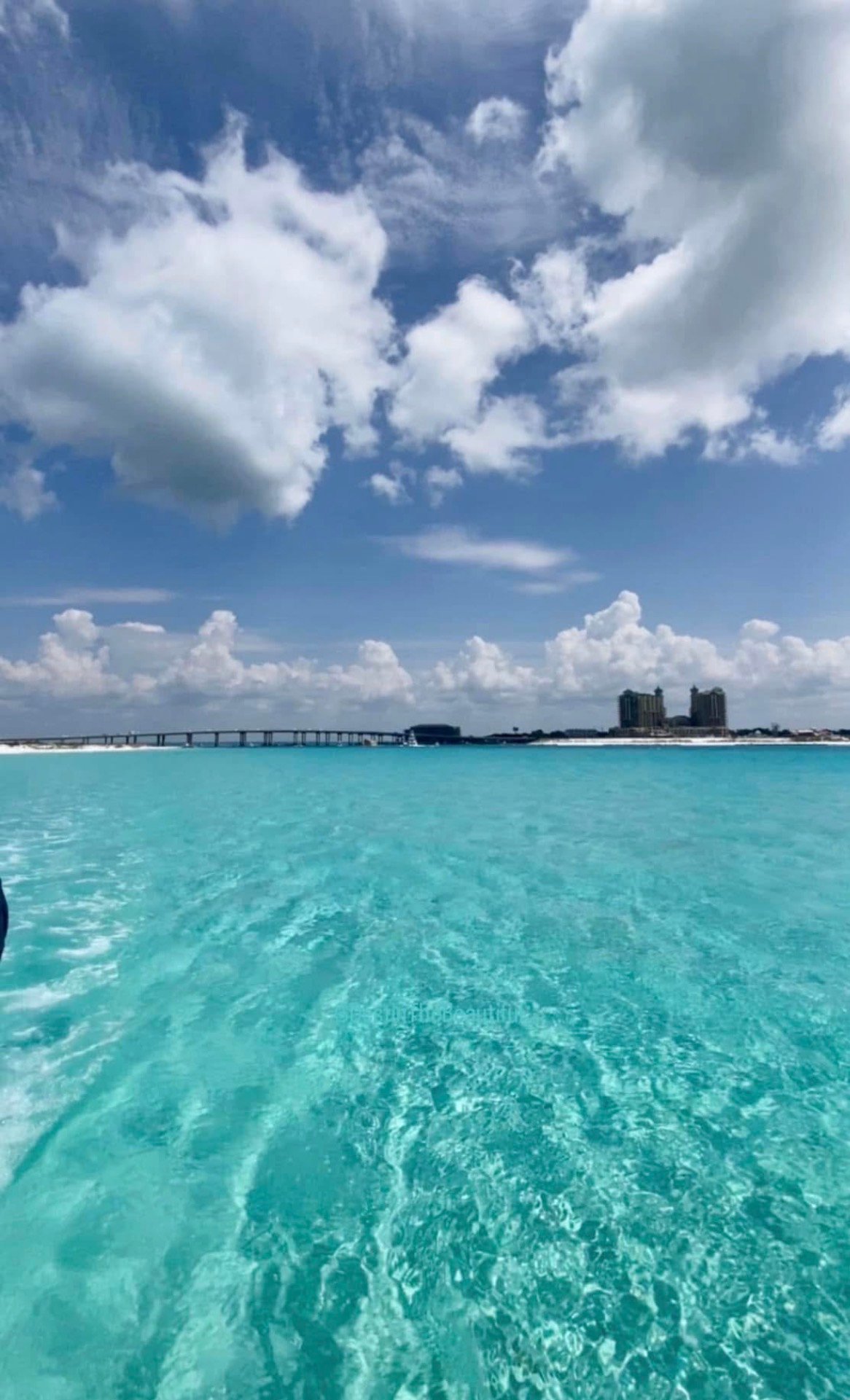 Turquoise coastal water beneath billowing clouds, with a bridge and distant beachfront buildings.