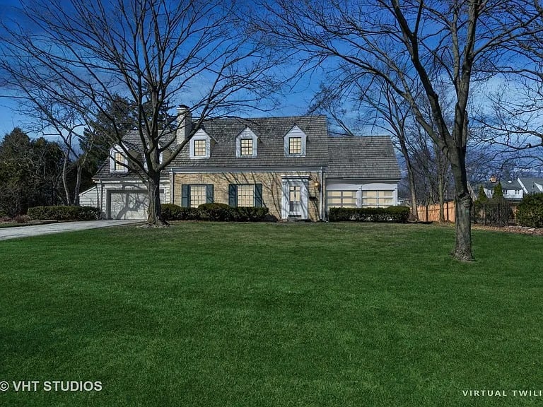 Beige two-story house with dormer windows, an attached garage, and a broad green lawn.