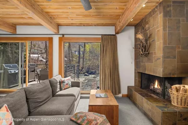 Living room with a stone fireplace, exposed timber ceiling, and glass doors to a deck.