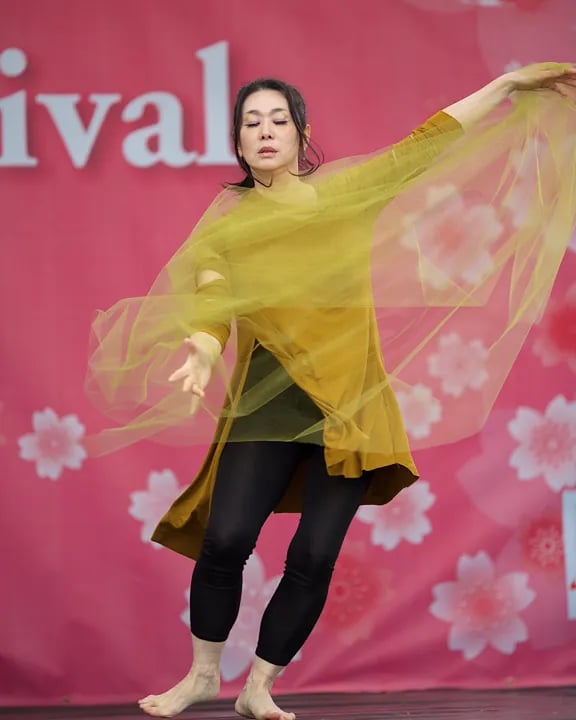A barefoot dancer in a yellow top and black leggings performs on stage, swirling a sheer yellow fabric against a pink cherry blossom backdrop at a festival.