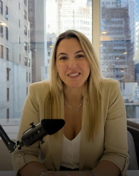 Woman in a beige blazer speaking into a desk microphone in front of city windows.