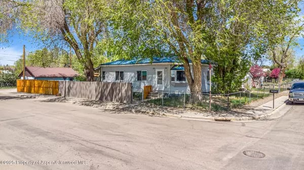 Single-story home with a blue roof beneath mature trees, viewed from a residential street corner.