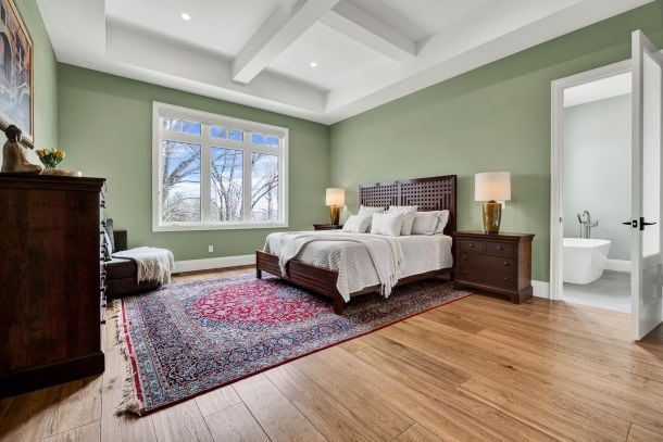 Staged bedroom with sage green walls, a coffered ceiling, patterned rug, and adjoining bathroom.