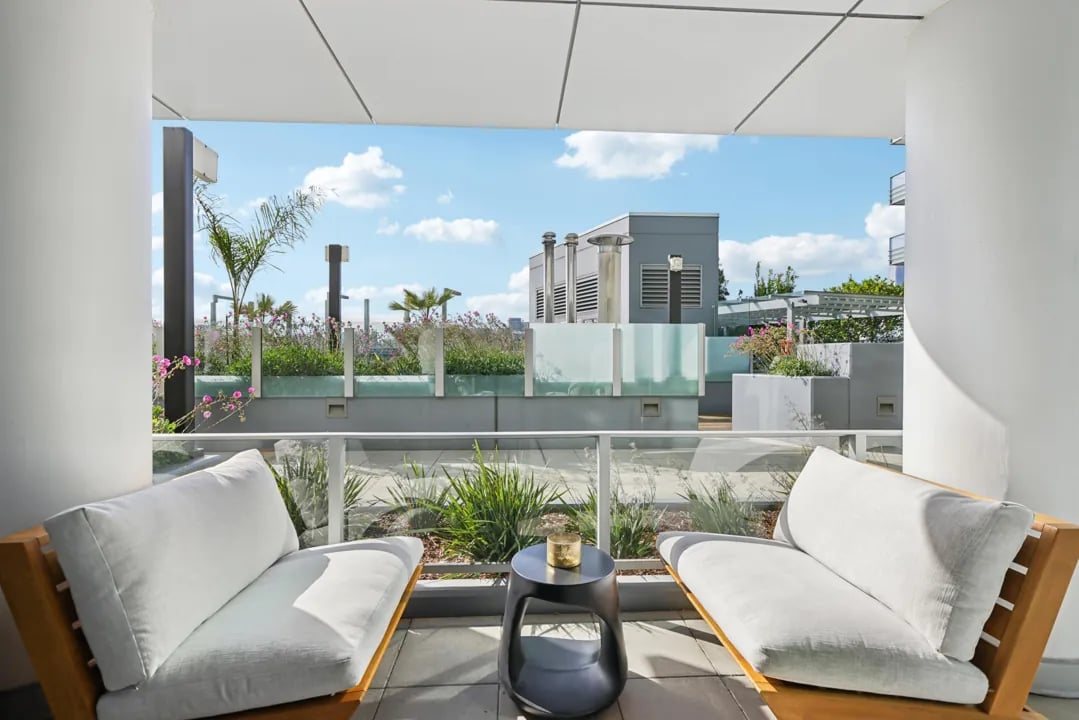 Two white outdoor lounge chairs and a small black table on a balcony with a view of an urban rooftop garden.