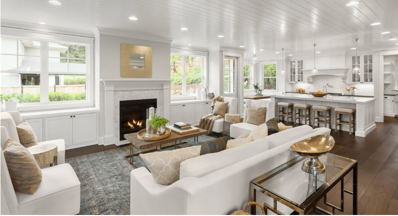 Open-plan living room and kitchen with a lit fireplace, white cabinetry, and large windows.