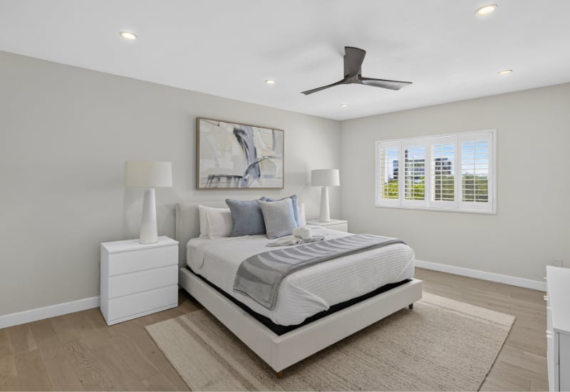 Staged bedroom with a white bed, gray bedding, abstract wall art, and shuttered windows.