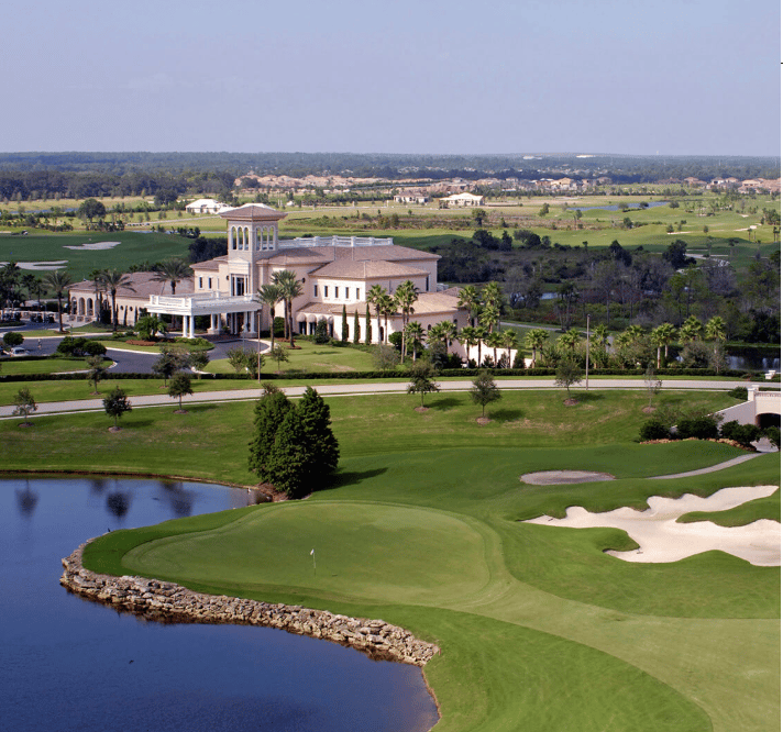 Golf course green beside a lake, with bunkers and a country club clubhouse in the distance.