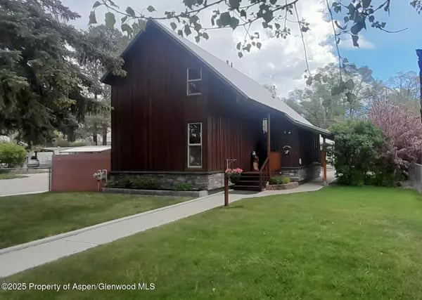 Dark red-brown home with a steep roof, stone foundation, and green lawn.