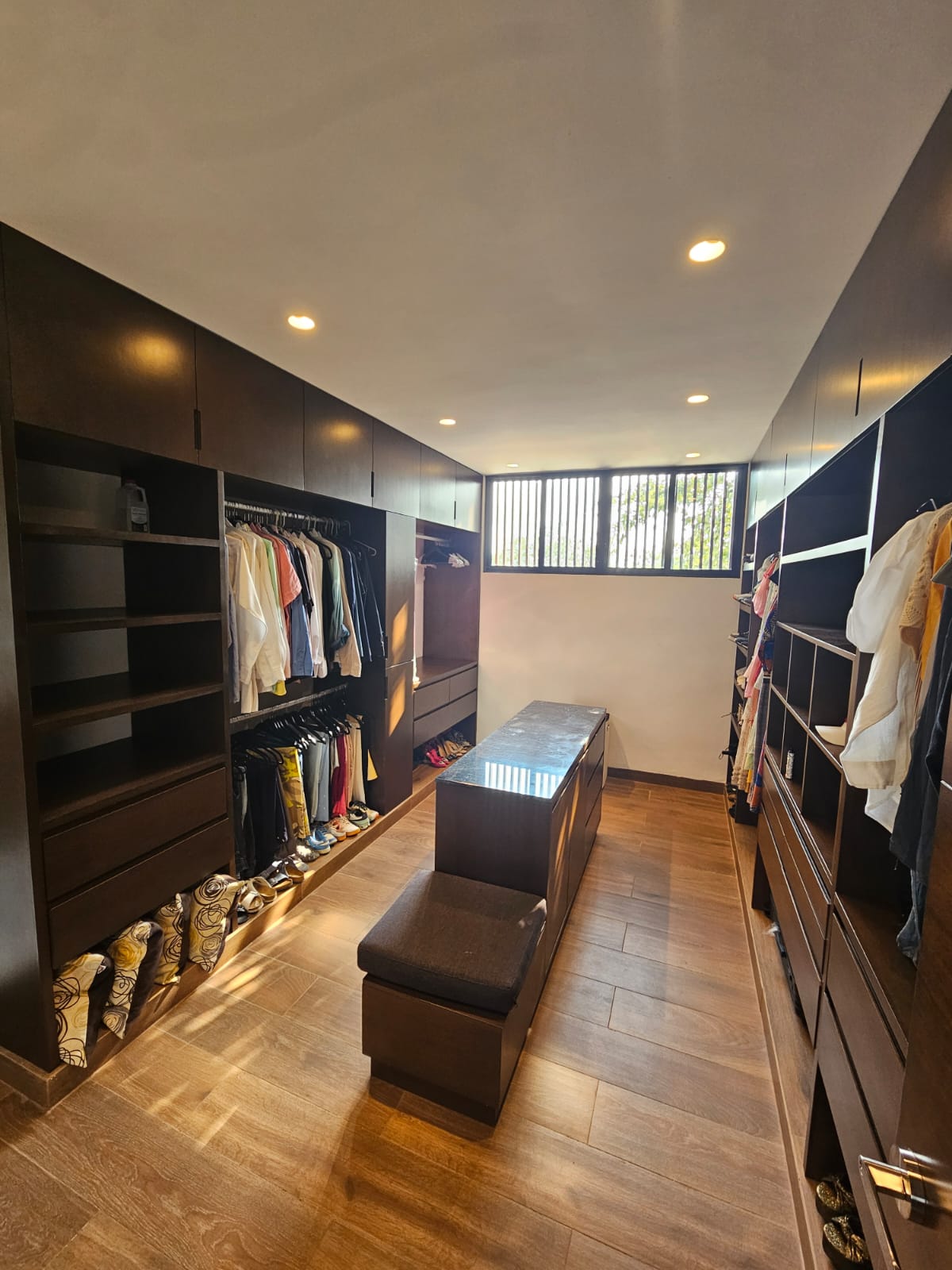 Walk-in closet with dark wood cabinetry, hanging clothes, and a central island and bench.