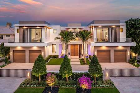 Modern two-story home with balconies, twin garage doors, palm trees, and a landscaped front garden at dusk.