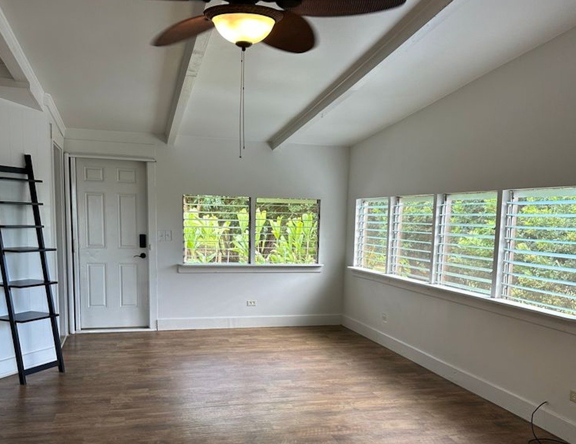 Empty room with vaulted ceiling, ceiling fan, wood-look flooring, and louvered windows facing tropical greenery.