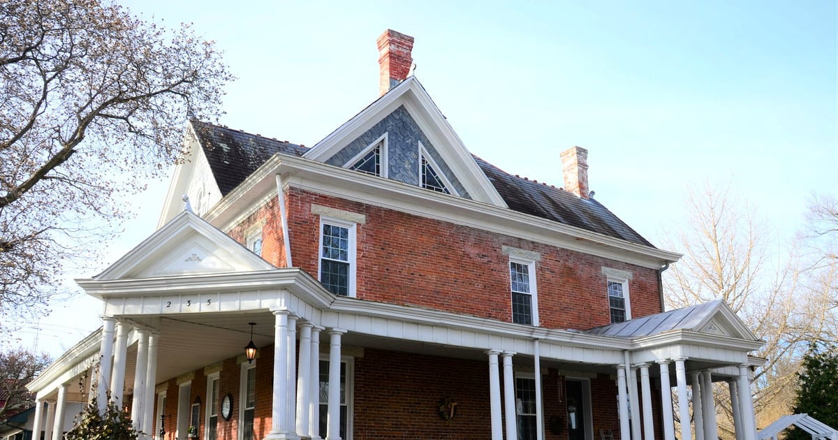 A large red brick two-story home with a white wrap-around porch on a clear day