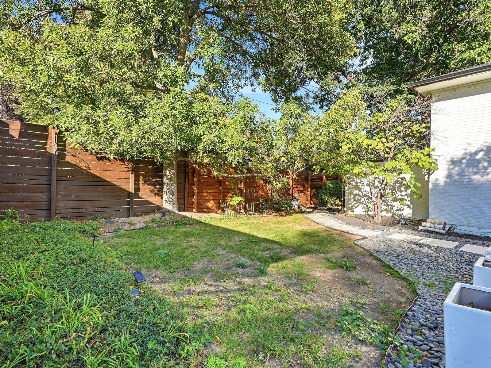 Backyard with mature trees, a wooden privacy fence, lawn, and pebble path beside the house.