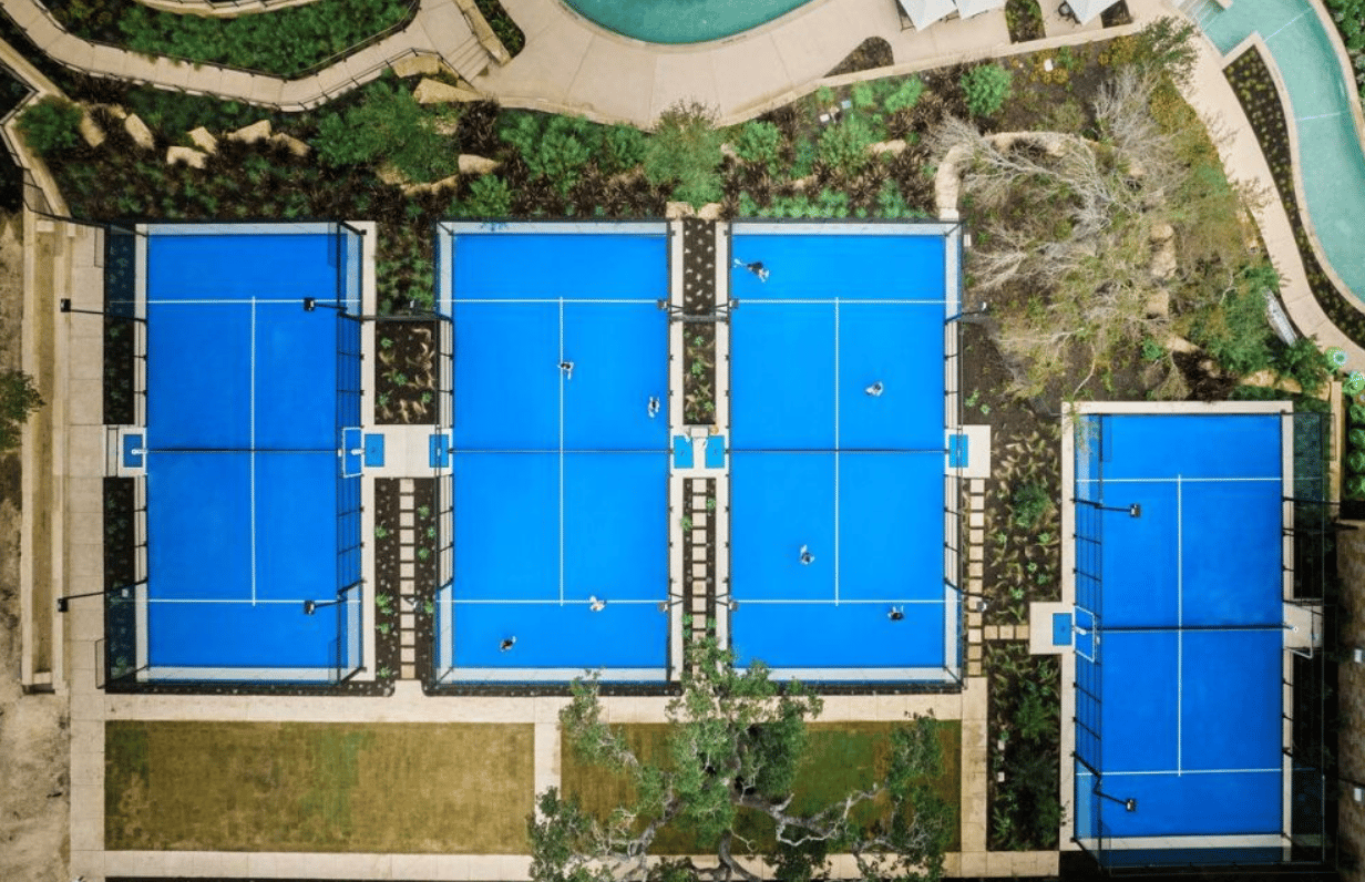 Four blue pickleball courts surrounded by landscaped paths, with a swimming pool nearby.