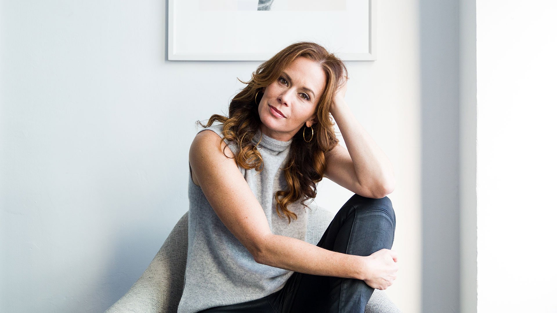 A woman with wavy hair sits in a chair, resting her head on her hand, wearing a gray sleeveless top and black pants.