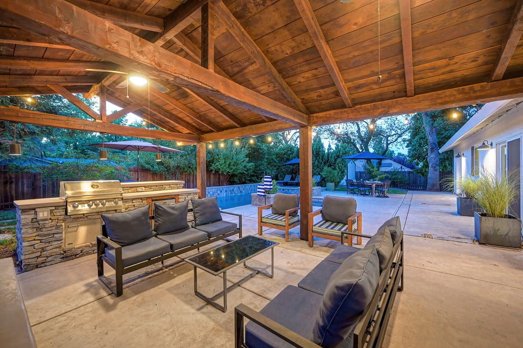 Covered patio with timber beams, cushioned seating, stone outdoor kitchen, and pool at dusk.