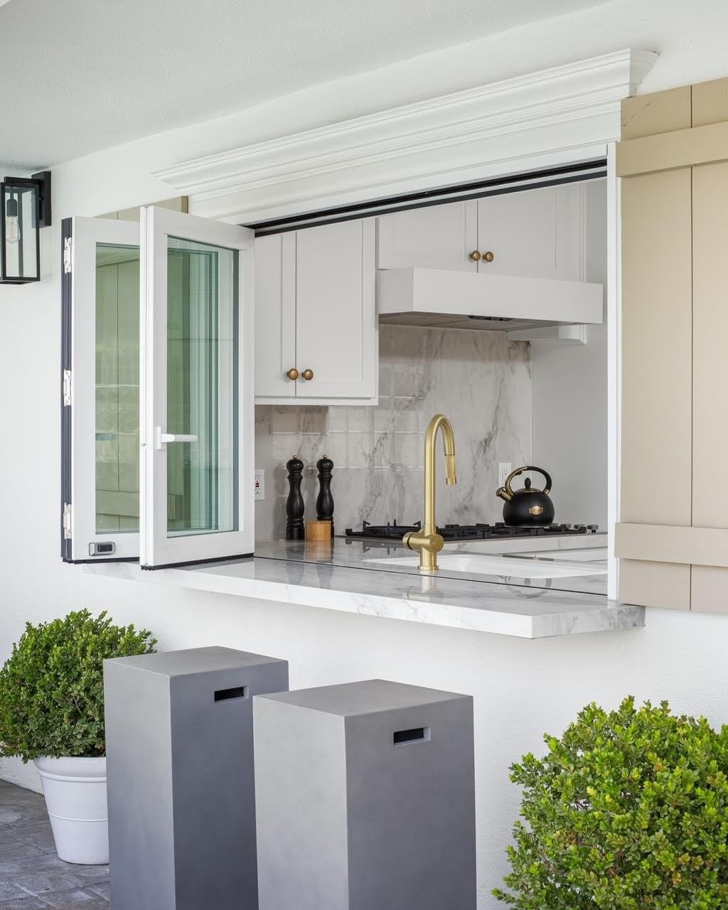  A bright outdoor kitchen pass-through window with a gold faucet, a black kettle, and two modern gray bar stools.